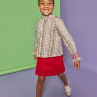 Model can be seen wearing a long sleeve green/red/white woven shirt that features button closures down the front, smocked red detail at sleeve cuffs, and ruffle detail at neckline. Paired back perfectly in our knit sparkly red skort with two pockets on the front to complete the look.