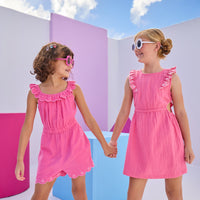 Model on the left can be seen wearing a hot pink gauze romper that has an elastic waistband, elastic collar, pockets, and a gorgeous embroidery detail at bottom of shorts for an elevated look. Model on the right can be seen wearing a hot pink dress in the same gauze fabric that has matching embroidery detail along the trim of sleeve and also features a cutout on each side of the dress for an elevated big girl look.