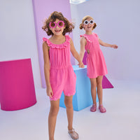 Model on the left can be seen wearing a hot pink gauze romper that has an elastic waistband, elastic collar, pockets, and a gorgeous embroidery detail at bottom of shorts for an elevated look. Model on the right can be seen wearing a hot pink dress in the same gauze fabric that has matching embroidery detail along the trim of sleeve and also features a cutout on each side of the dress for an elevated big girl look.