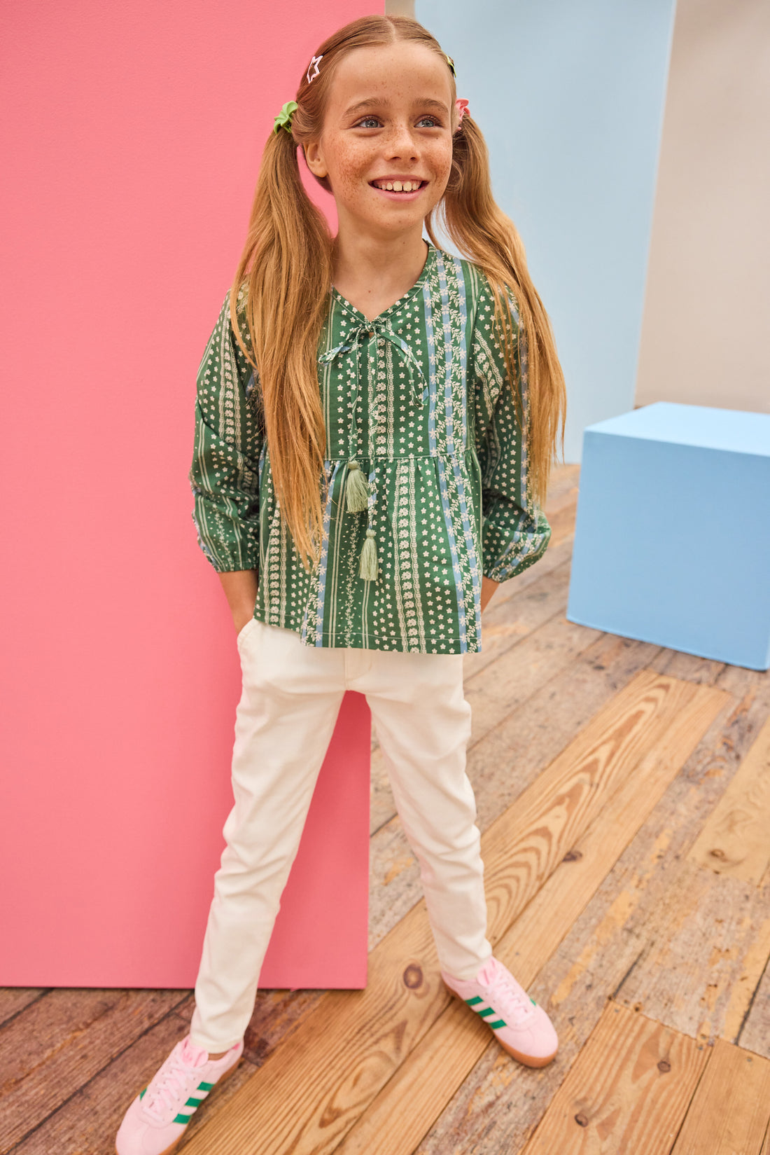 Young girl in a green patterned shirt and white pants standing on a wooden floor with colorful walls.