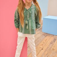 Young girl in a green patterned shirt and white pants standing on a wooden floor with colorful walls.