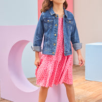 Young girl wearing a pink dress with a denim jacket and heart-shaped sunglasses in front of colorful geometric shapes.