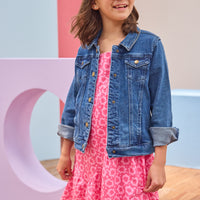 Young girl wearing a pink dress with a floral pattern and a blue denim jacket, standing against a colorful background.