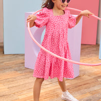 Young girl in a pink dress with a floral pattern, holding a pink hula hoop on a wooden floor.