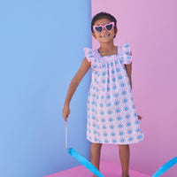 Model can be seen wearing a woven dress in a fun blue/pink/red print. Dress features a square neckline with pink ribbon detail across edge, pink pom pom detail at end of sleeves, and and angel sleeve detail to elevate the look.