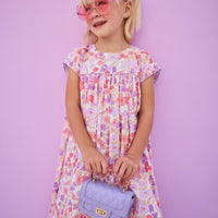 Girls/tween woven dress in a lavender/salmon floral print with matching lavender piping across bust and sleeves. Dress features button closures along back and made of a lightweight material that is lined for a secure but comfy fit.