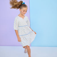Model can be seen wearing a woven 3/4 sleeve length dress that has button closure along back in a blue and green floral print. Dress features a blue ric rac detail on the top and bottom tier of the dress to elevate the look.