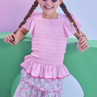 Model can be seen wearing a woven top in a light pink color that features smocking along the whole body of the shirt, angel sleeves, and hot pink trim at bottom to elevate the look. Top pairs back perfectly with tiered skort in a pink floral print to complete the look.