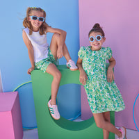 Tween model on left can be seen wearing shirred shorts in a green/light blue floral print. Shorts feature smocking along waist and scallop edges to elevate the look. Paired back perfectly with a white knit tank top with fun lace features across chest to complete the look. Tween model on right can be seen in a knit dress in the same print for a matching moment.