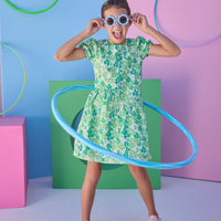 Tween model can be seen wearing a woven dress in a blue and green floral print. Dress features an asymmetrical design, elastic cuffs at sleeves, and button closure along back for an elevated but secure fit.