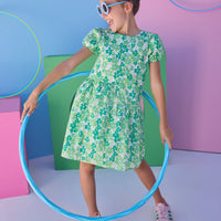 Tween model can be seen wearing a woven dress in a blue and green floral print. Dress features an asymmetrical design, elastic cuffs at sleeves, and button closure along back for an elevated but secure fit.