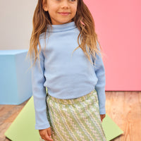 Model can be seen wearing a soft light blue ribbed turtleneck paired back with a beautiful green skort that features a silver metallic and white pattern to elevate the look.