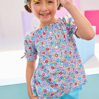 Girls/tween model can be seen wearing woven lilac/red/blue floral pattern top that features an elastic neckline, button closures along front, and faux ties at elastic cuffed sleeves. Top pairs back perfectly with knit turquoise shorts to complete the look.