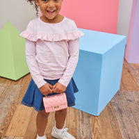 Tween girl seen wearing a pale pink and white stripe ruffle long sleeve t-shirt paired back with a pleated denim skort to elevate the look.