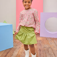 Model can be seen wearing long sleeve woven top in a pink/green floral pattern. Top features a button closure at back and a pleating feature at bust for an elevated look. Top pairs back perfectly with a corduroy skort in the matching green to complete the look.