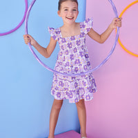 Model can be seen wearing a woven dress in a purple floral print. Dress features smocking detail across bust and slight angel sleeves to elevate the look.