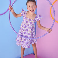 Model can be seen wearing a woven dress in a purple floral print. Dress features smocking detail across bust and slight angel sleeves to elevate the look.