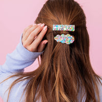 Person with long brown hair wearing colorful hair clips on a pink background