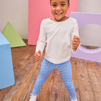 Child wearing a white long-sleeve shirt and blue patterned leggings standing on a wooden floor with colorful geometric shapes in the background.