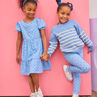 Two young girls in matching blue outfits standing against a pink wall.