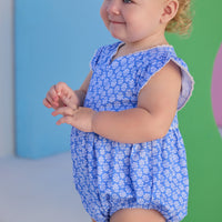 Baby model can be seen wearing a knit bubble in a blue and white floral print. Bubble features white ric rac details on edge of angel sleeves, button closure along back, and stride for an easy put on/pull off access.