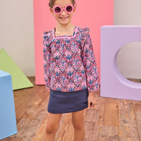 Model can be seen wearing a woven long sleeve top in a navy/pink/red floral print, paired back with a quilted navy skort. Top features a square neckline, with a ribbon detail along neckline and slight angel sleeves to elevate the look.