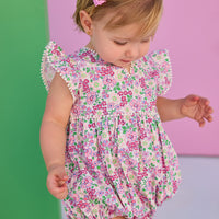 Baby model can be seen wearing a woven bubble in a pink and green floral print. Bubble features white pom pom details on edge of angel sleeves, button closure along back, and stride for an easy put on/pull off access.