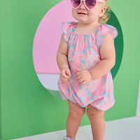 Baby girl seen in a woven pink and blue floral bubble. Bubble features scallop sleeves, elastic bust line, and button snap closure at stride.