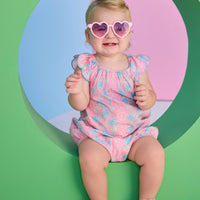 Baby girl seen in a woven pink and blue floral bubble. Bubble features scallop sleeves, elastic bust line, and button snap closure at stride.