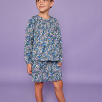 Model can be seen wearing a long sleeve woven dress in a blue/purple/white floral print. Top features ruching across the bust and elastic at the sleeves.