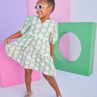 Model can be seen wearing a woven 3/4 sleeve length dress that has button closure along back in a green and salmon pink floral print. Dress features a white ric rac detail on the top and bottom tier of the dress to elevate the look.