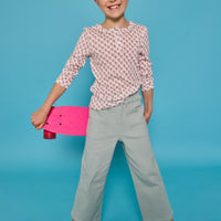 Model can be seen wearing a long sleeve knit tee in an orange/green floral print. Top features three button closure at neckline and a ruffle collar for an elevated look. Top pairs back perfectly with our light green pants that feature two pockets and elastic waistband for easy pull on access.