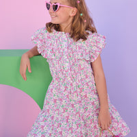 Model can be seen wearing a woven dress in a pink/green floral print. Dress features angel sleeves, button closure down the front, and tier details to elevate the look.