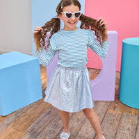 Young girl in a striped shirt and metallic skirt standing in front of colorful geometric shapes on a wooden floor.