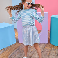 Young girl playing with her hair against a colorful geometric background wearing a blue metallic skirt and blue and white striped long sleeve top.