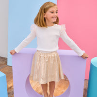 Young girl in a white ribbed long sleeve top gold metallic skirt standing on a purple platform with a colorful background.
