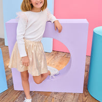 Young girl in a white ribbed long sleeve top gold metallic skirt standing on a purple platform with a colorful background.
