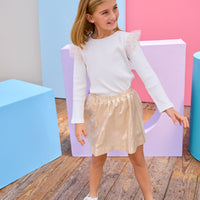 Young girl in a white ribbed long sleeve top gold metallic skirt standing on a purple platform with a colorful background.