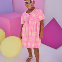 Model can be seen wearing a knit terry material dress in bubblegum pink with yellow flowers. Dress features v-neckline with tassel ties to elevate the look.