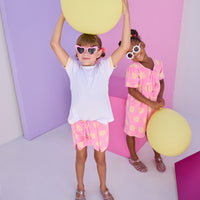 Model can be seen wearing knit terry shorts in a bubblegum pink/yellow floral print. Shorts feature tassel tie detail along with pom pom details along front and side of shorts. Shorts pair back perfectly with white tee with the same yellow/pink colored ric rac detail on the cuffs of sleeves.