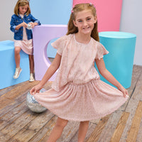 Young girl in a pink crinkle top and skort posing in front of colorful cylindrical objects.