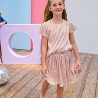 Young girl in a pink crinkle top and skort standing on a wooden floor with colorful geometric shapes in the background.