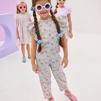 Girls/Tween model in front can be seen wearing a woven jumpsuit that features ruffle detail along the front/sleeves, pockets, and a front tie with an open back. Jumpsuit has a beautiful pale yellow/pink/green/blue floral pattern and is a lightweight woven material for ultimate comfort.