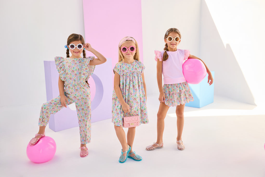 Girls/Tween model on left can be seen wearing a woven jumpsuit that features ruffle detail along the front/sleeves, pockets, and a front tie with an open back. Jumpsuit has a beautiful pale yellow/pink/green/blue floral pattern and is a lightweight woven material for ultimate comfort. Model in the center can be seen in the same floral in a woven dress, and model on right can be seen in the same print with our tiered skort paired back with a light pink eyelet tank.