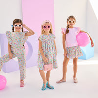 Girls/Tween model on left can be seen wearing a woven jumpsuit that features ruffle detail along the front/sleeves, pockets, and a front tie with an open back. Jumpsuit has a beautiful pale yellow/pink/green/blue floral pattern and is a lightweight woven material for ultimate comfort. Model in the center can be seen in the same floral in a woven dress, and model on right can be seen in the same print with our tiered skort paired back with a light pink eyelet tank.