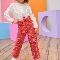 Tween model can be seen wearing an elevated white long sleeve shirt featuring ruffle detail and white scallop detail across the entirety of the shirt paired back with a red and purple floral print pant. Pants feature a belt and zipper/button clasp for a secure fit.
