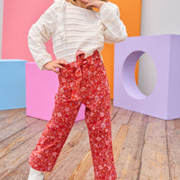 Tween model can be seen wearing an elevated white long sleeve shirt featuring ruffle detail and white scallop detail across the entirety of the shirt paired back with a red and purple floral print pant. Pants feature a belt and zipper/button clasp for a secure fit.