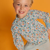 Model can be seen wearing an orange/green/teal floral print woven shirt that features buttons down the front with ruffled detail along neckline and smocking detail at sleeves for an elevated look. Paired back perfectly with our light green pants with elastic waistband and pockets in the front.