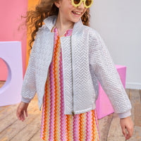 Young girl wearing a colorful dress and a silver quilted jacket with yellow sunglasses, standing on a wooden floor with a colorful abstract background.