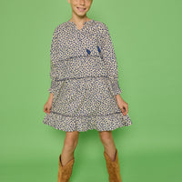 Model can be seen wearing a long sleeve woven dress in a blue/cream floral print. Dress features tier detail along body, elastic cuffs at sleeves, and v-neckline with tassel detail to complete the look.
